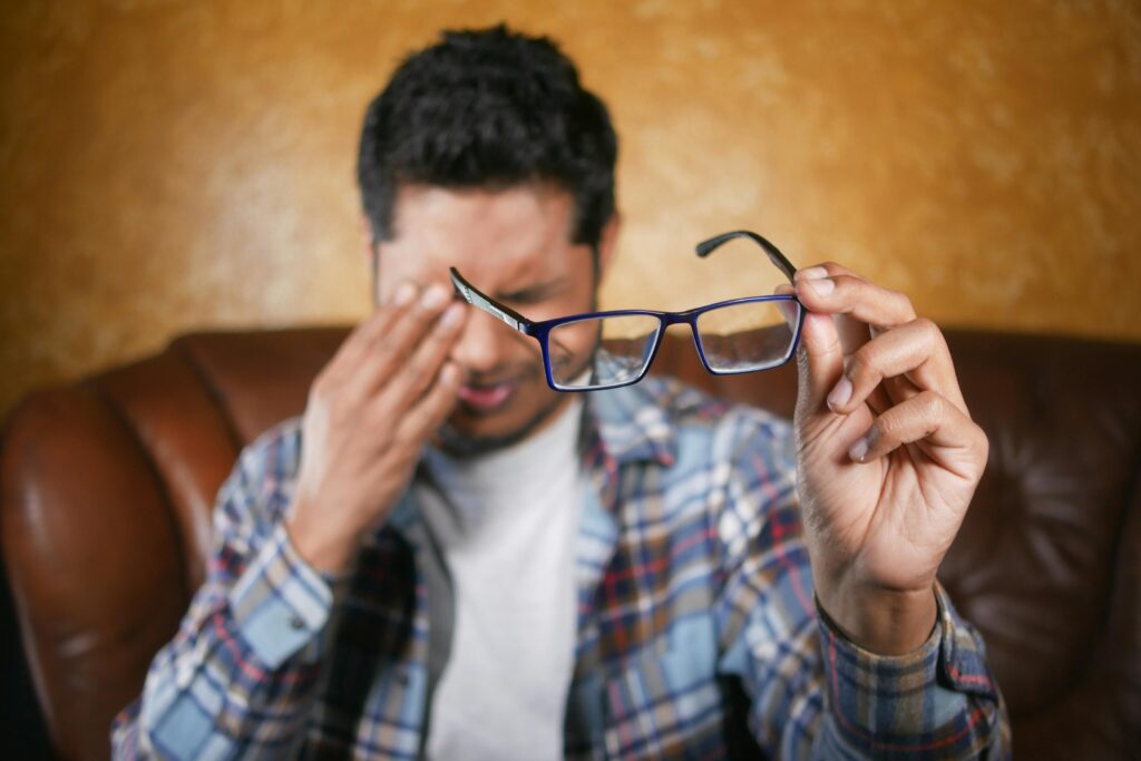 A man in plaid shirt holding eyeglasses indoors. Focus on eyewear with neutral background.